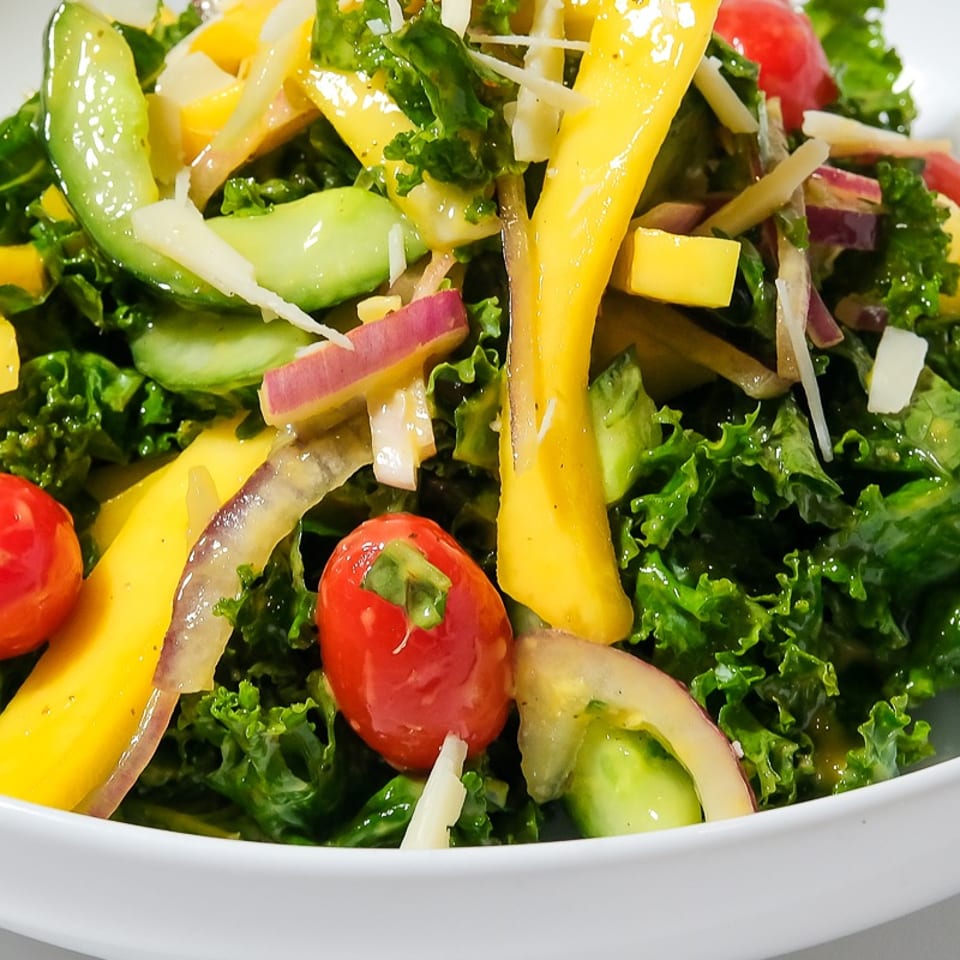 Fresh kale salad with mango, cucumber, cherry tomatoes, red onion, and shaved cheese in a white bowl.