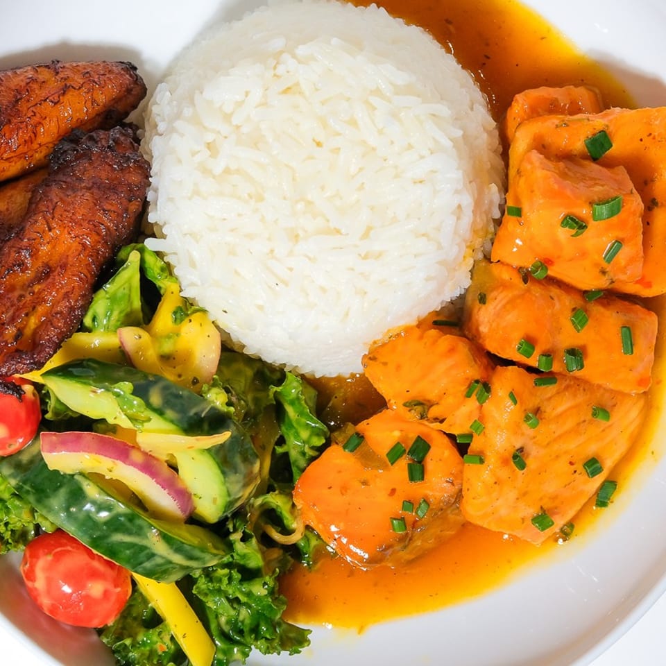 Plate of white rice with orange-sauced salmon, fried plantains, and mixed green salad with tomatoes and cucumber.
